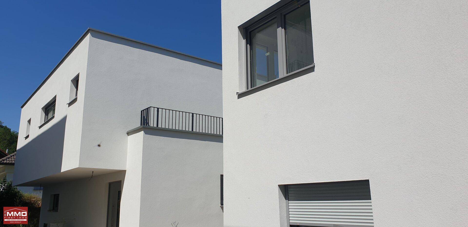 Moderne weiße Fassade eines Gebäudes mit klaren Linien, Flachdach und einem Fenster mit Rollladen.