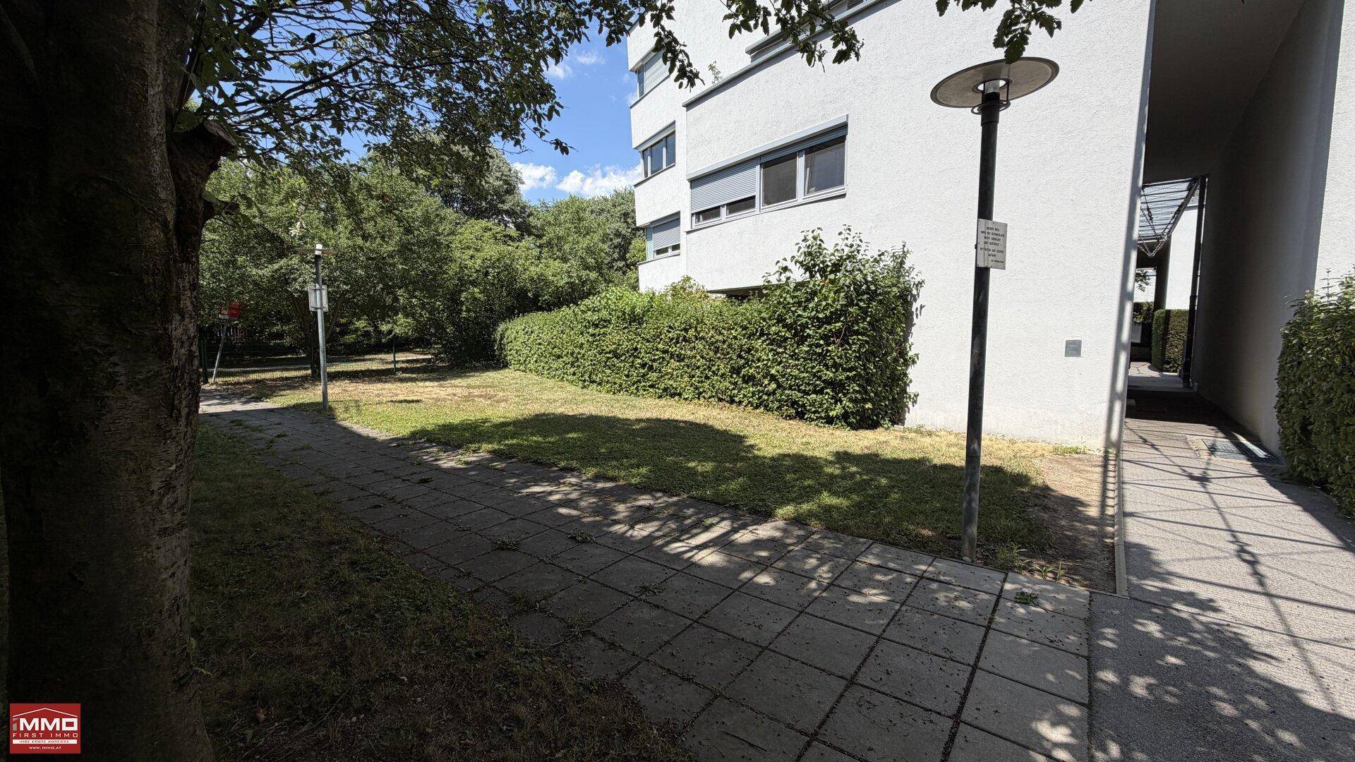 Gepflegte Grünfläche mit Rasen und Bäumen neben einem Wohngebäude an einem sonnigen Tag.