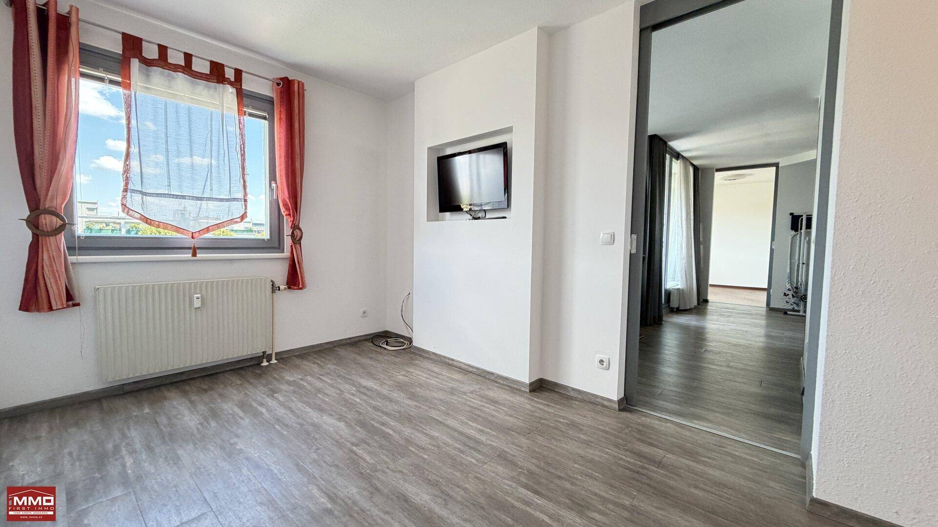 Heller Raum mit Laminatboden, weißen Wänden und einem an der Wand montierten Fernseher.