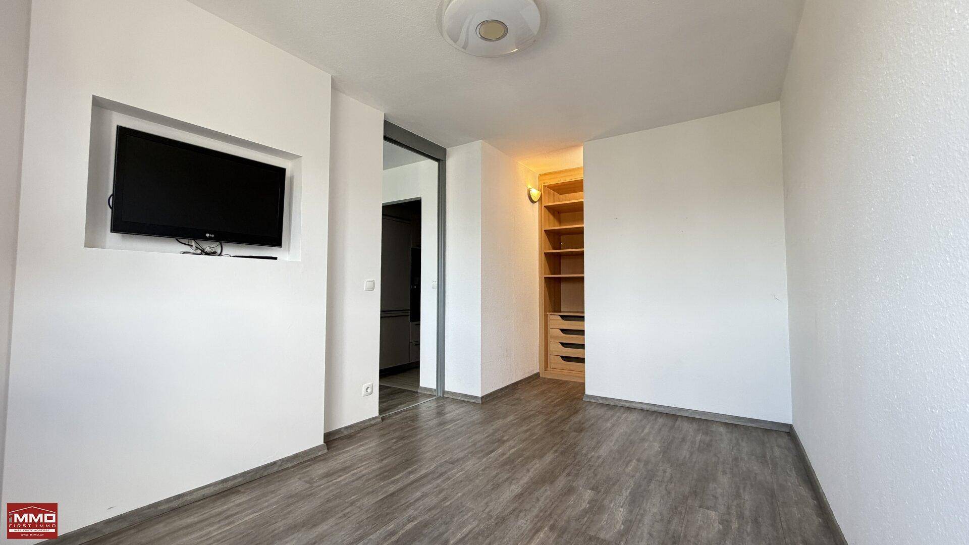 Raum mit Laminatboden, Wandfernseher und Blick in einen begehbaren Kleiderschrank.