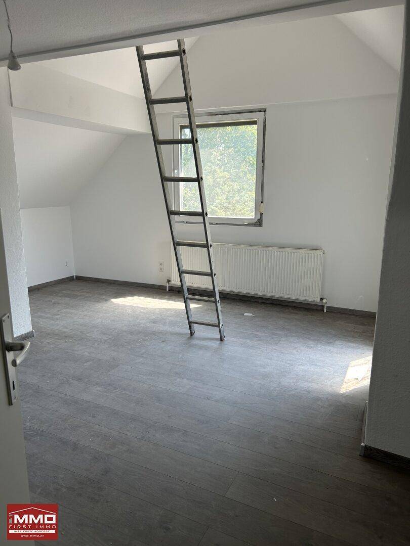 Helles Zimmer im Dachgeschoss mit Laminatboden und Fenster, bietet Zugang zum Dachboden.