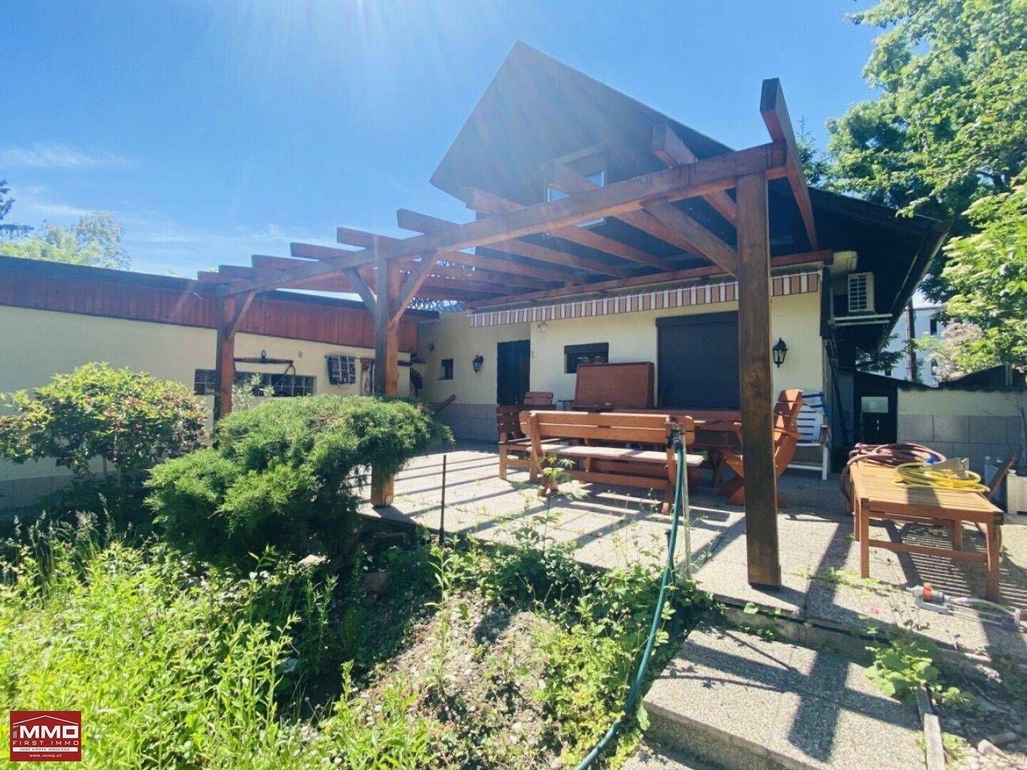 Geräumige Terrasse mit Holzmöbeln unter einer Pergola, umgeben von üppigem Grün.