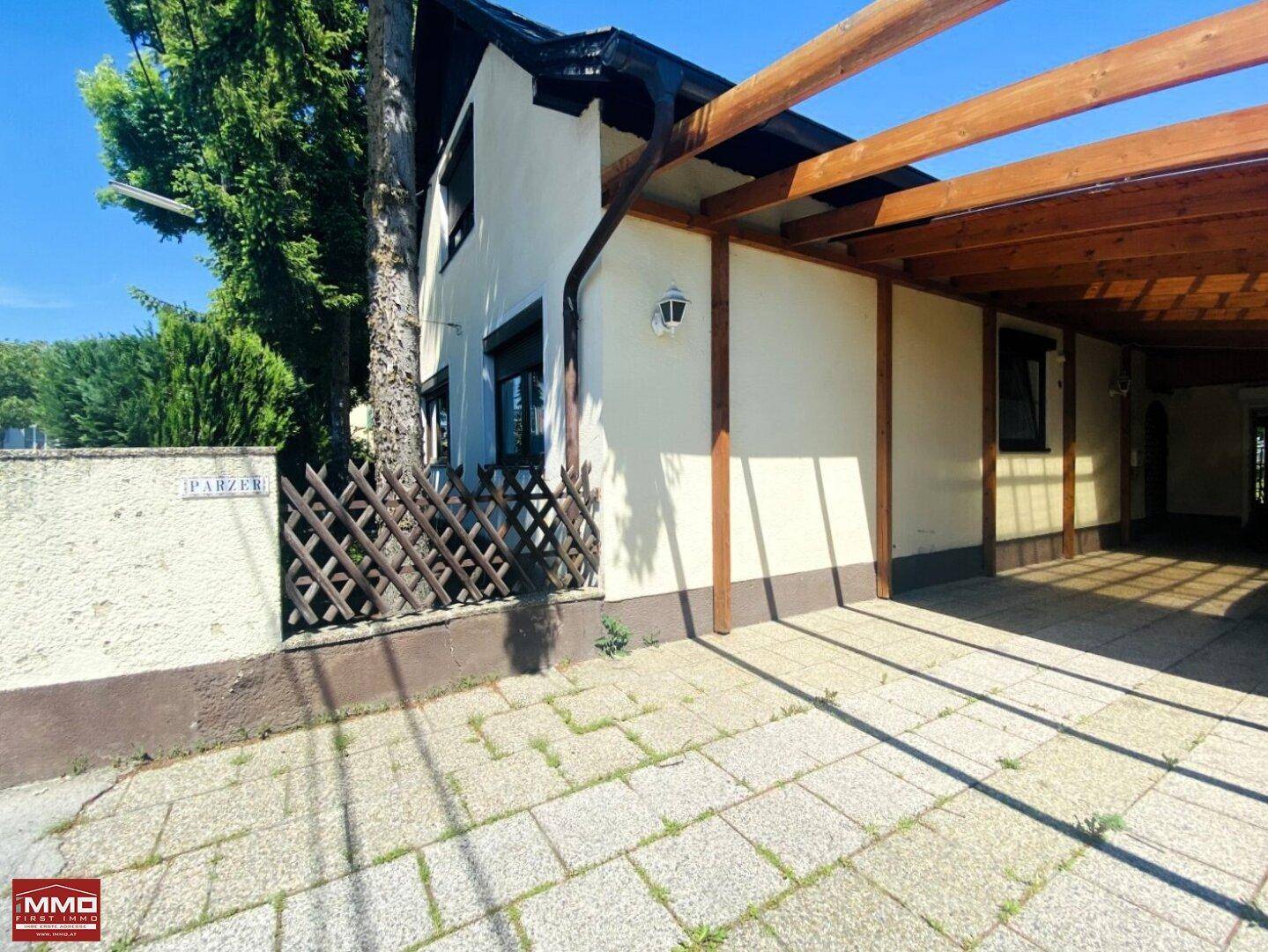Seitenansicht des Hauses mit Holzcarport und gepflastertem Bereich an einem sonnigen Tag.