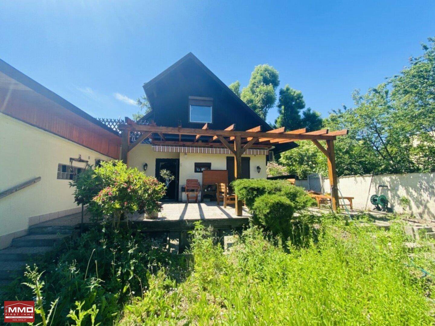 Hinteransicht des Hauses mit großer Terrasse unter einer Holzpergola und angrenzendem Garten.