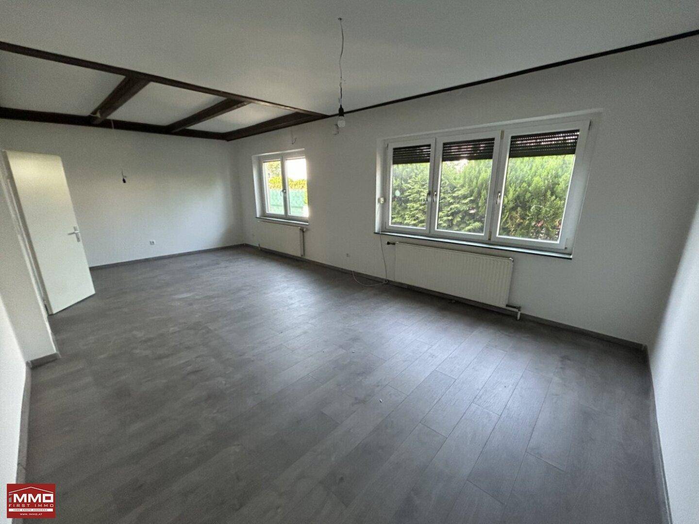 Geräumiges Wohnzimmer mit Laminatboden, sichtbaren Deckenbalken und großen Fenstern.