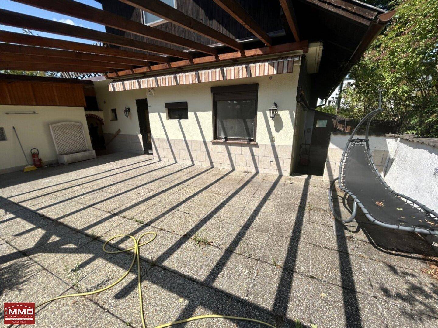 Gepflasterte Terrasse mit Schattenwurf der Pergola und einem Trampolin im Hintergrund.