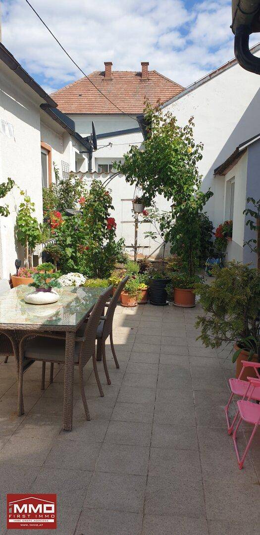 Gepflasterte Terrasse mit großem Esstisch und Stühlen, umgeben von Pflanzen und Gebäuden.