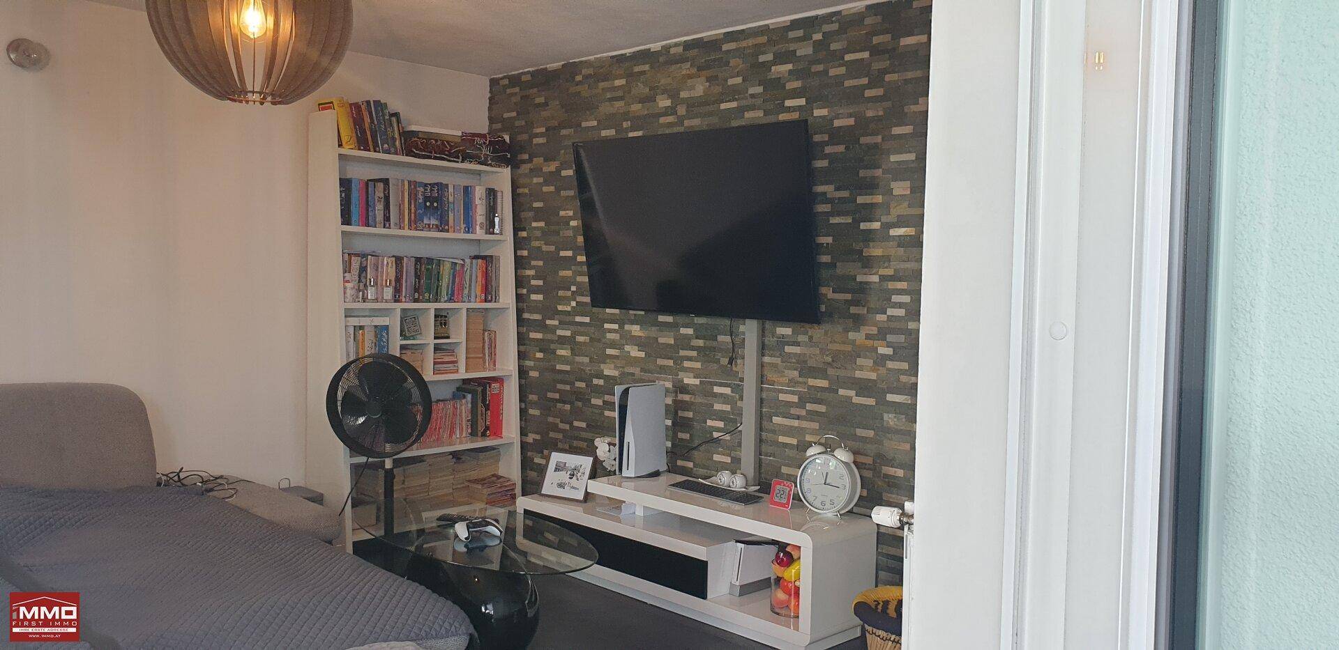 Wohnzimmer mit Fernseher an einer Steinwand, Bücherregal und gemütlichem Sofa.
