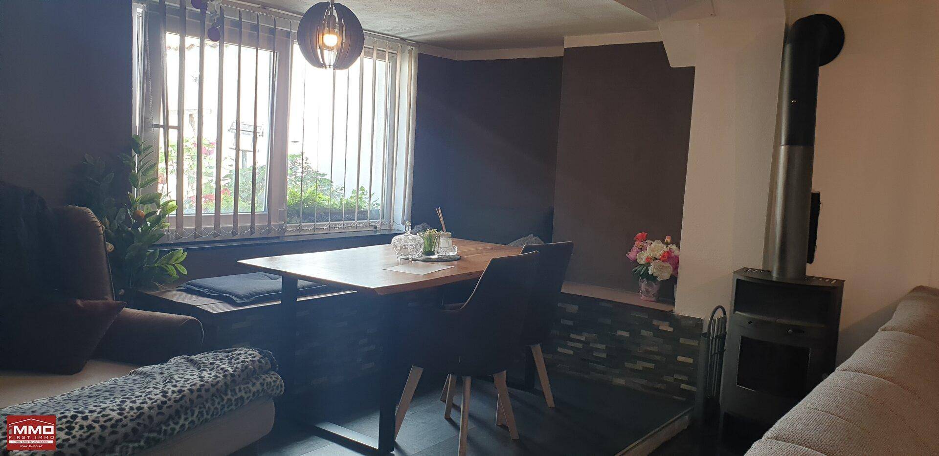 Gemütlicher Wohn- und Essbereich mit Holztisch, Sofa und einem Holzofen neben dem Fenster.