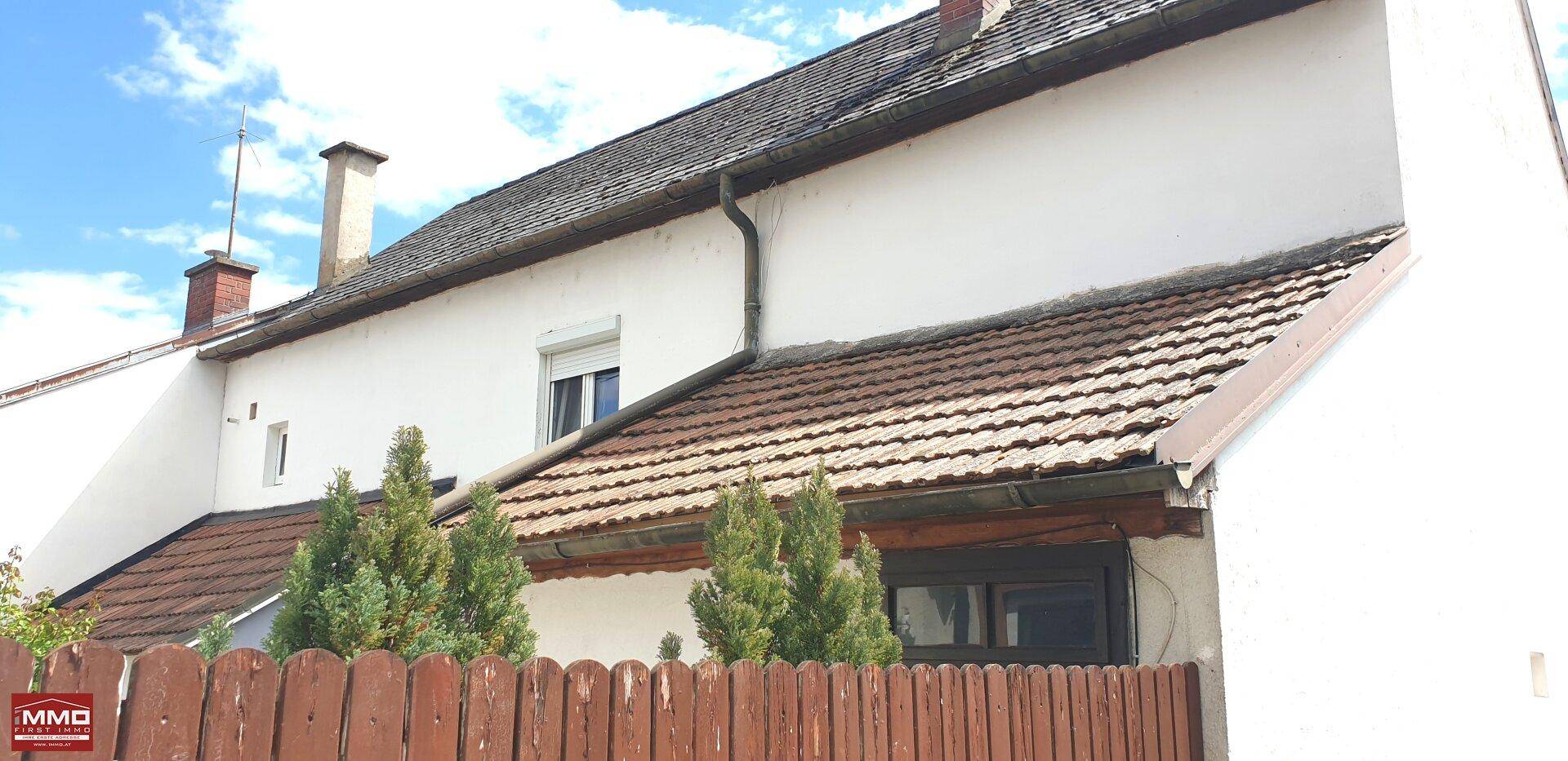 Seitenansicht eines weißen Hauses mit Ziegeldach, Rollläden und einem Holzzaun im Vordergrund.
