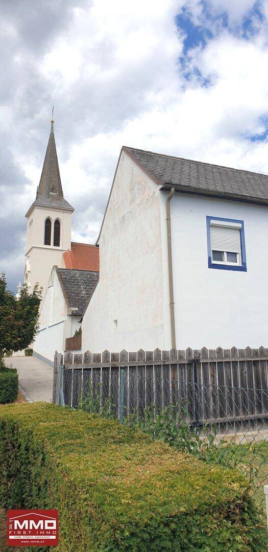 Weißes Wohnhaus mit blauen Fensterrahmen und Rollläden neben einer Kirche, umgeben von Hecken.