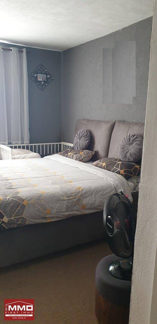 Schlafzimmer mit Doppelbett, Babybett und grauen Wänden, ausgestattet mit einem Ventilator.