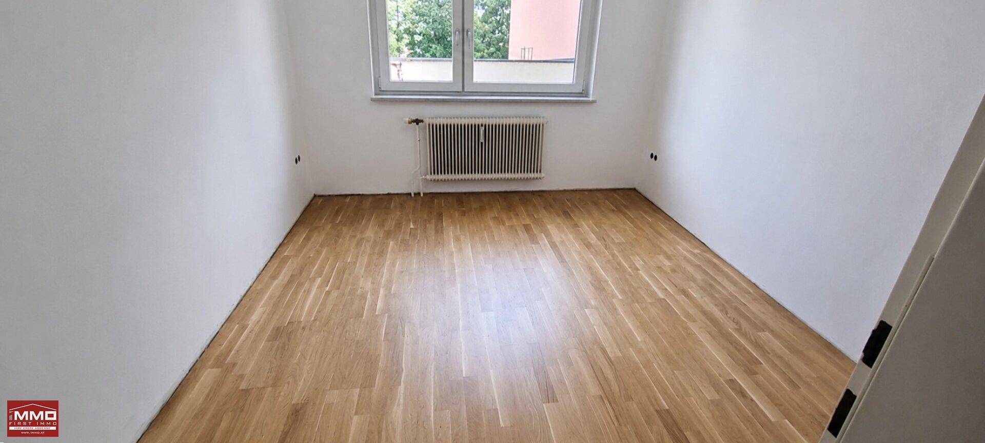 Leeres Zimmer mit hellem Holzfußboden und einem Fenster, das Tageslicht hereinlässt.