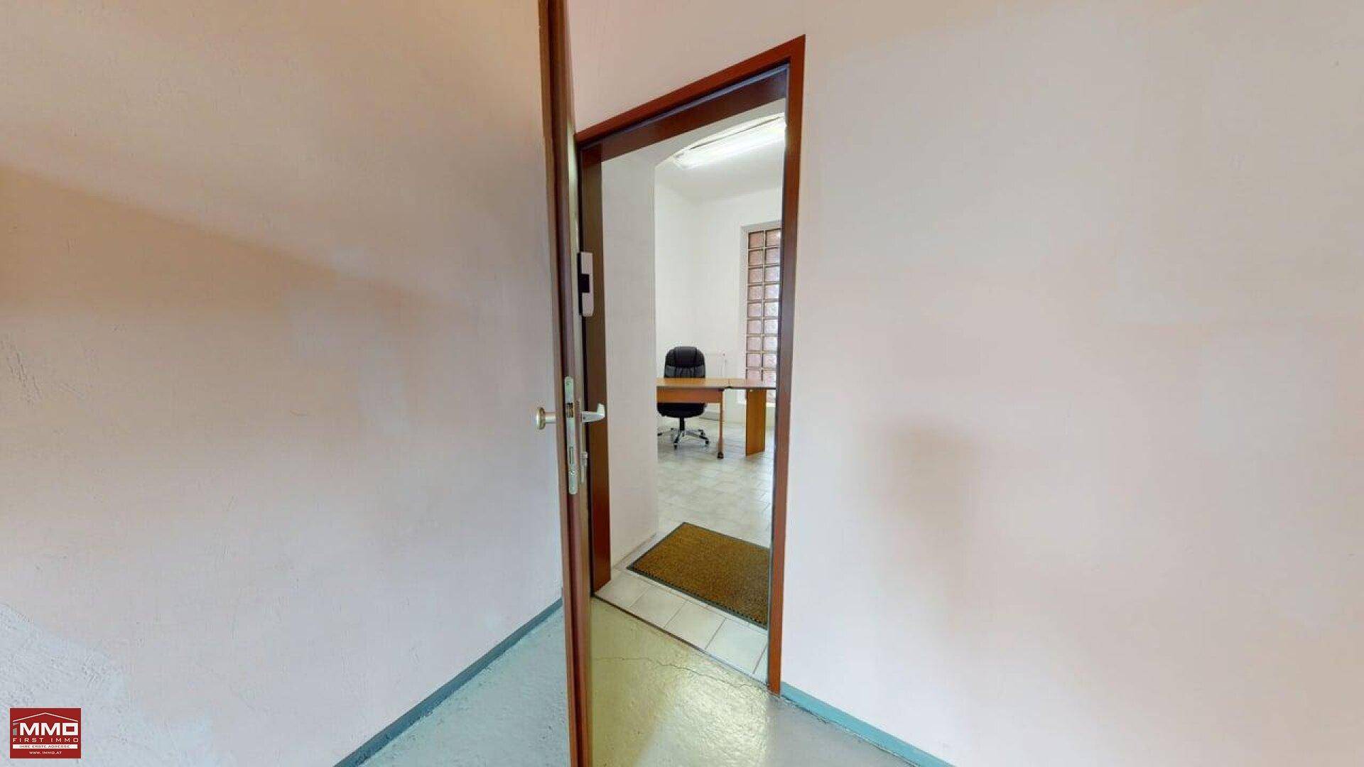 Blick durch eine offene Tür in ein Büro mit Schreibtisch und einem Fenster aus Glasbausteinen.