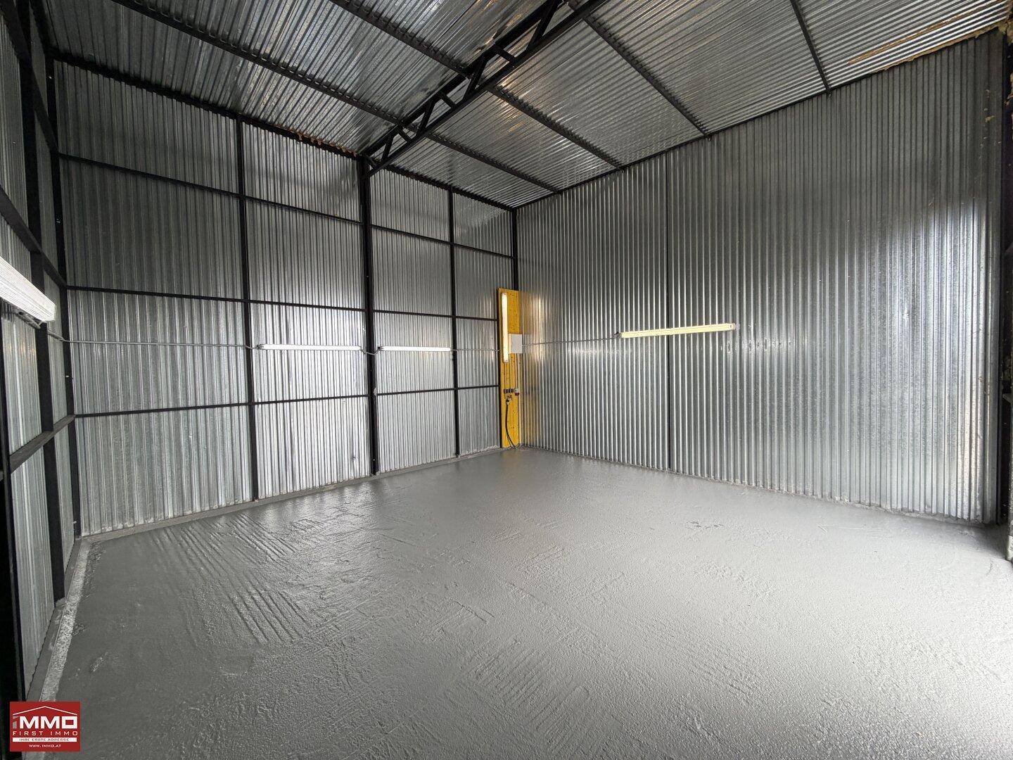 Weitläufige Lagerhalle mit Wellblechverkleidung, Betonboden und einer einzelnen Leuchtstoffröhre an der Decke.