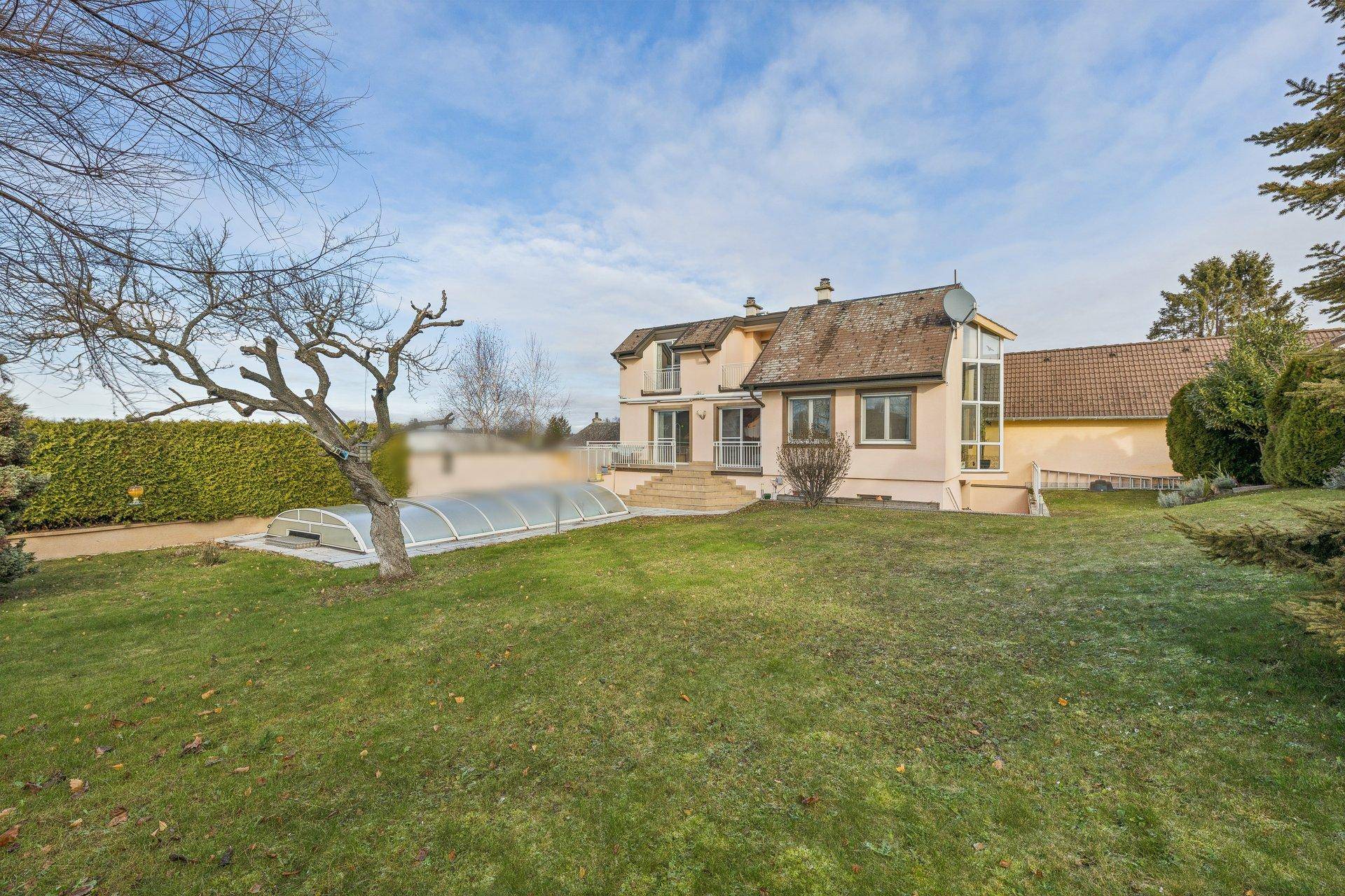 Hinteransicht des Hauses mit großem Rasen, umgeben von Bäumen und einem überdachten Poolbereich.