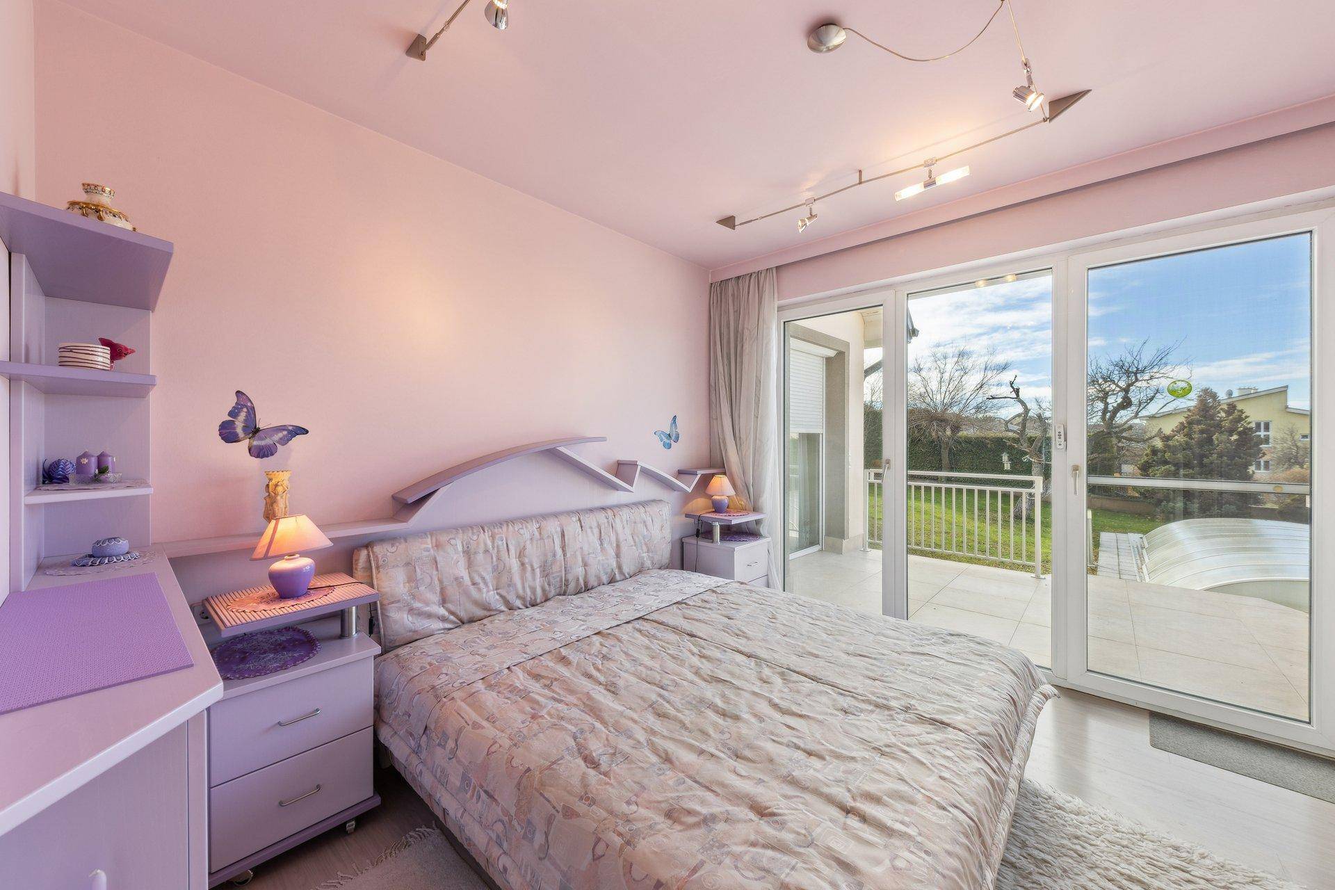 Schlafzimmer mit lila Dekoration, Zugang zu einem Balkon und Blick auf den Gartenbereich.