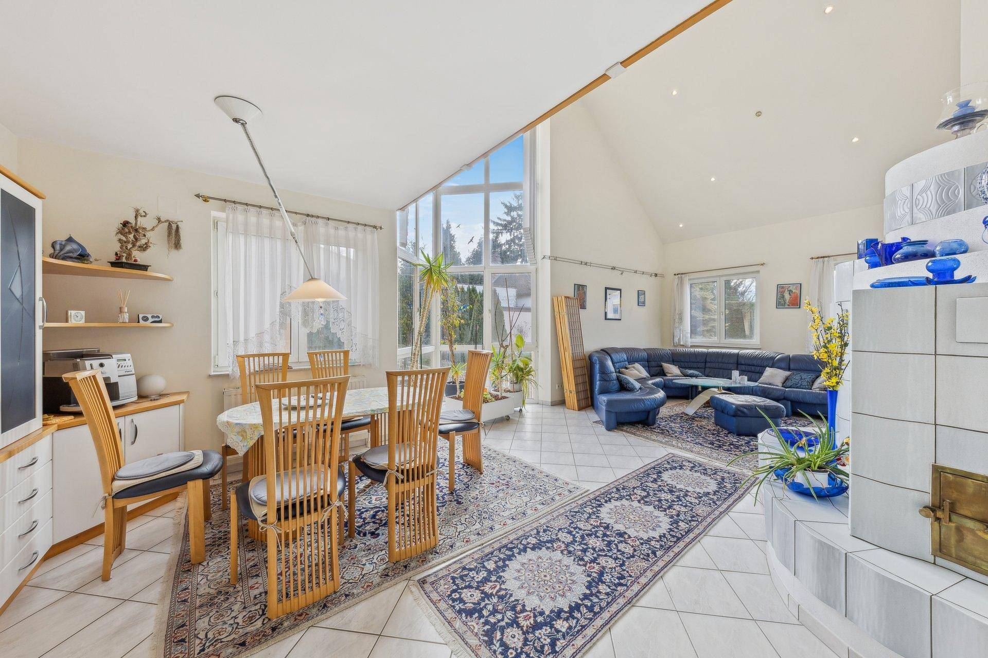 Großzügiger Wohn- und Essbereich mit Fliesenboden, Kachelofen und hohen Fenstern, die viel Licht hereinlassen.