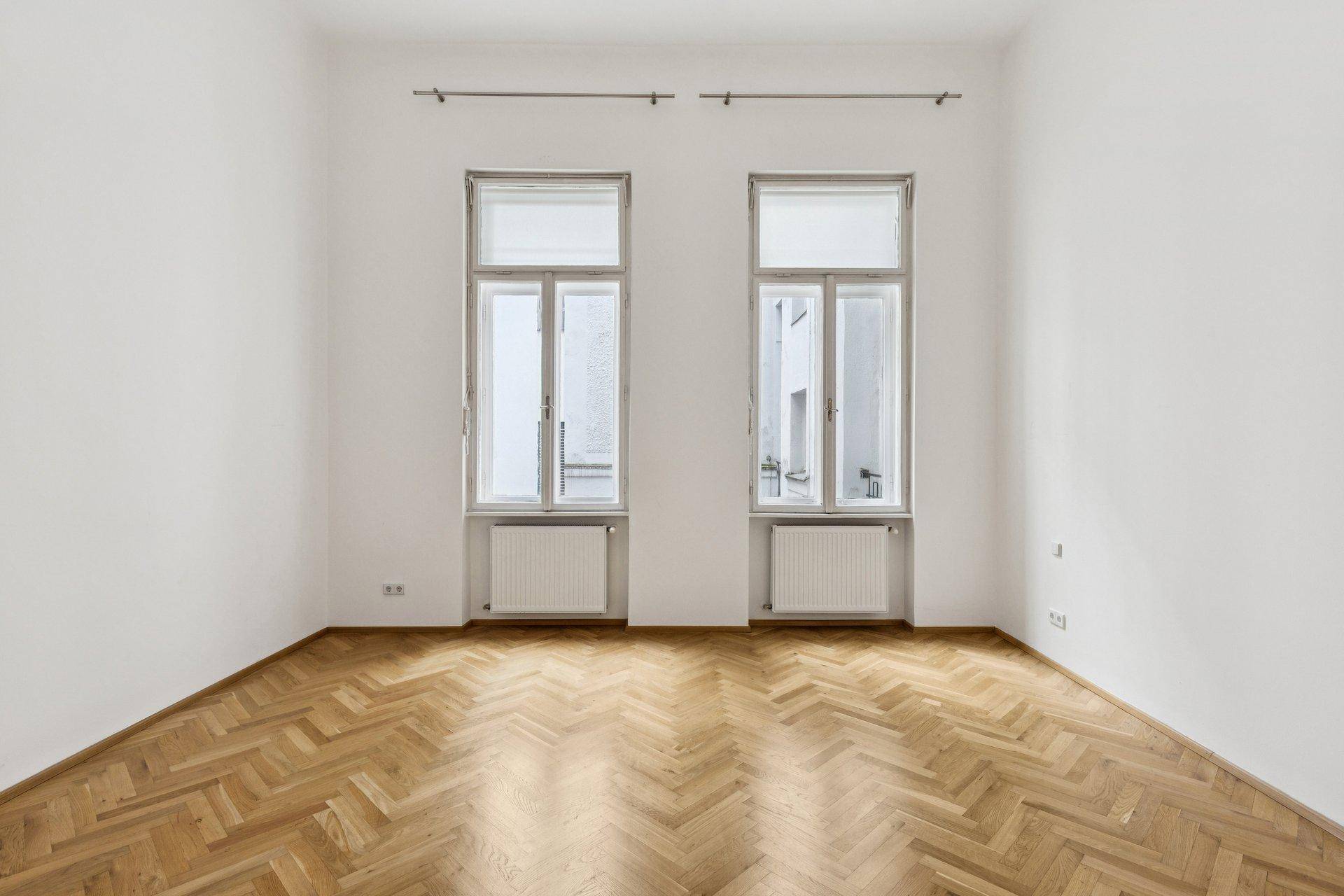 Helles Zimmer mit zwei hohen Fenstern und klassischem Fischgrätparkett.