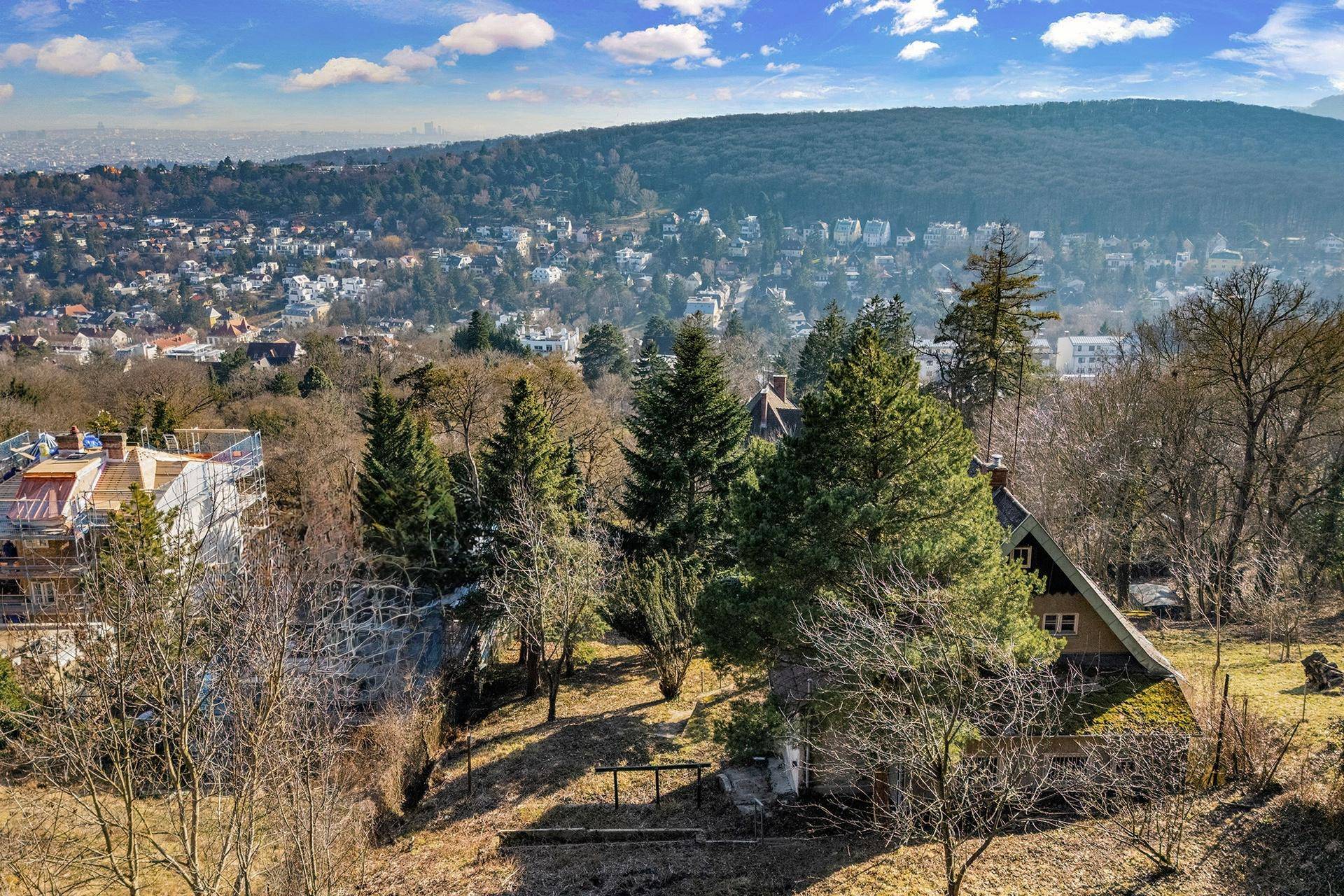 Haus in Hanglage, umgeben von hohen Bäumen und Blick auf die umliegende Hügellandschaft.