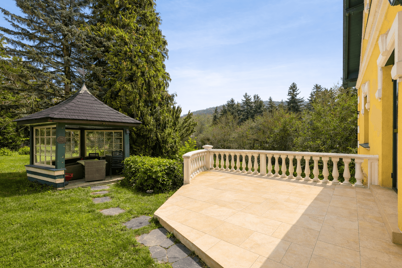 Große gepflasterte Terrasse mit Balustrade und Gartenpavillon, umgeben von Bäumen.