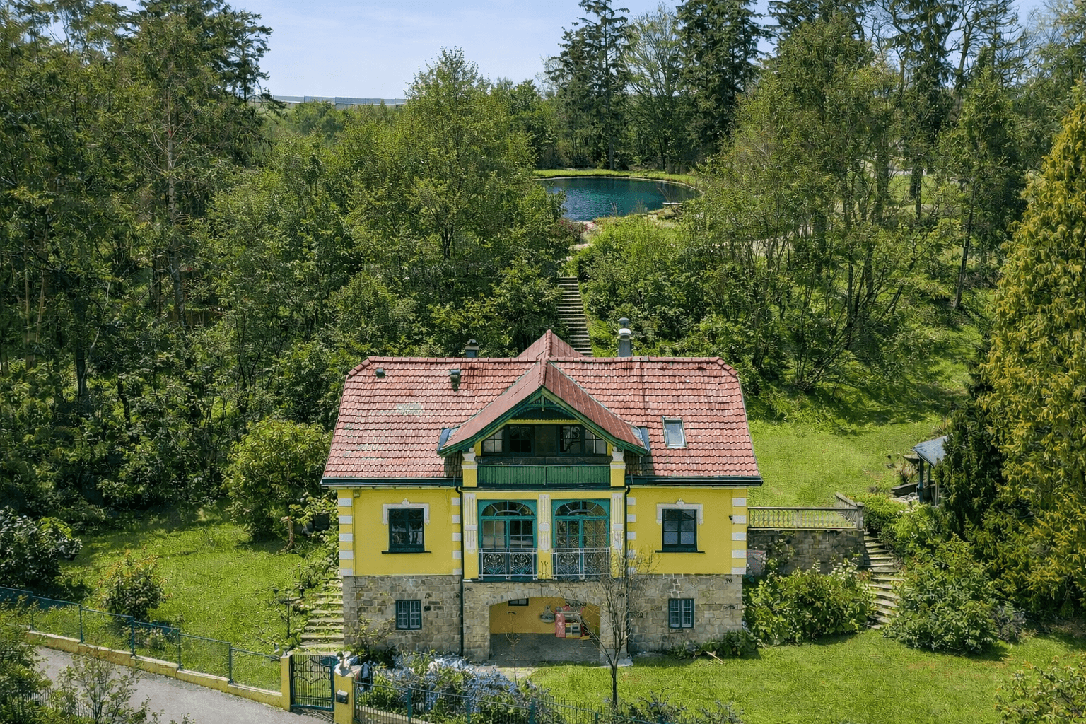 Historisches Gebäude mit rotem Ziegeldach und gelber Fassade, umgeben von viel Grün und einem Teich.