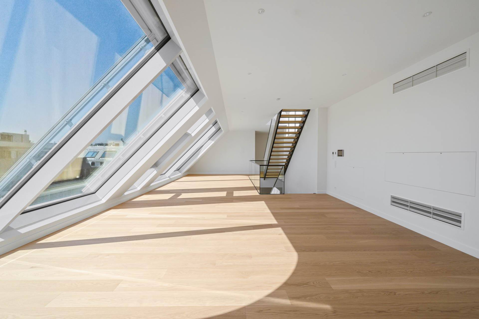 Geräumiger, heller Raum mit Holzboden, großen Fenstern und Treppe im Hintergrund.