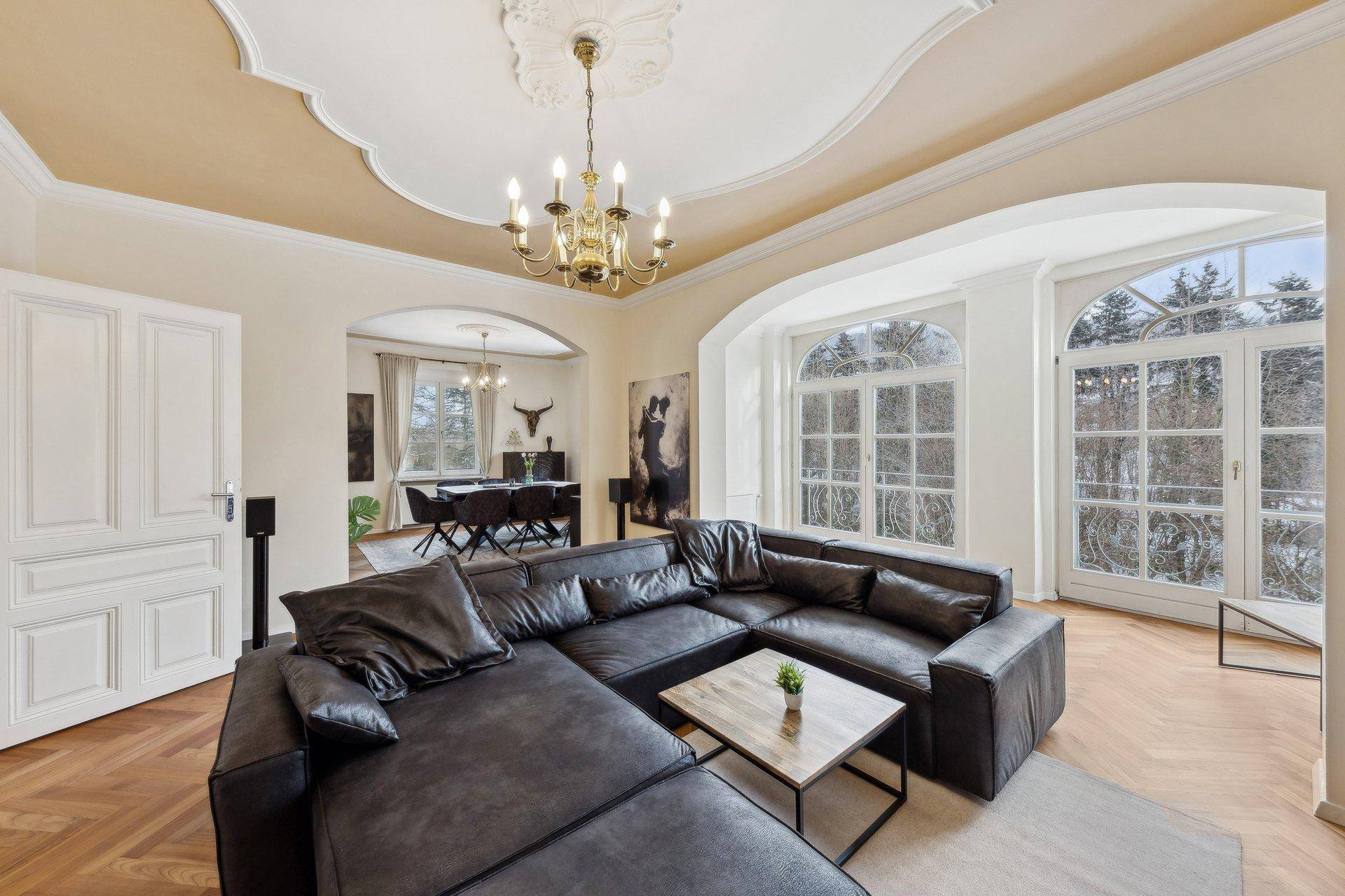 Elegantes Wohnzimmer mit Stuckdecke, Fischgrätparkett und großen Fenstern mit Blick ins Grüne.