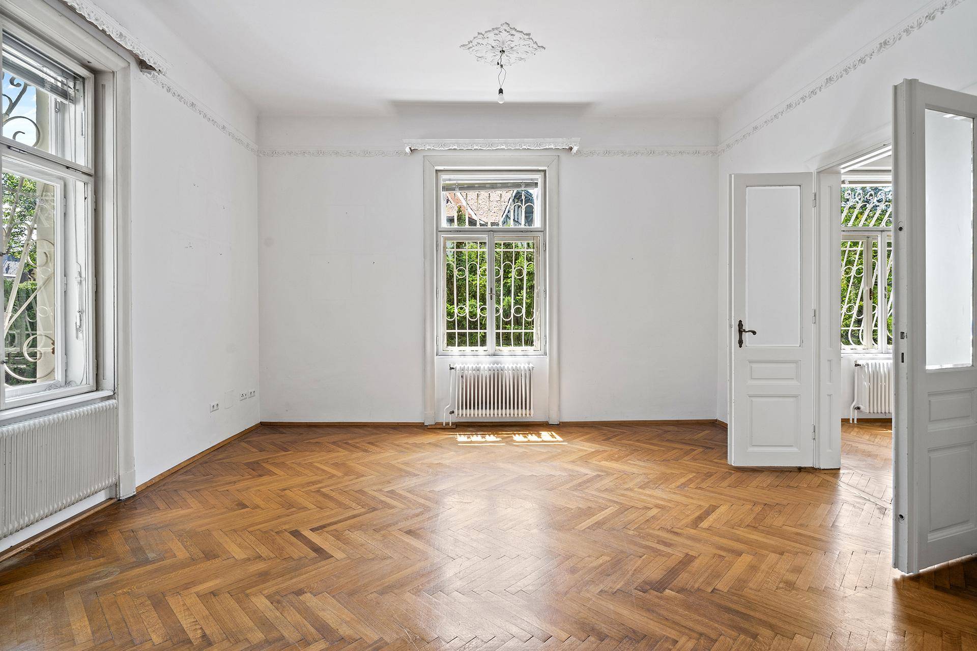 Leeres Wohnzimmer mit Fischgrätparkett, hohen Decken und Stuckverzierungen.