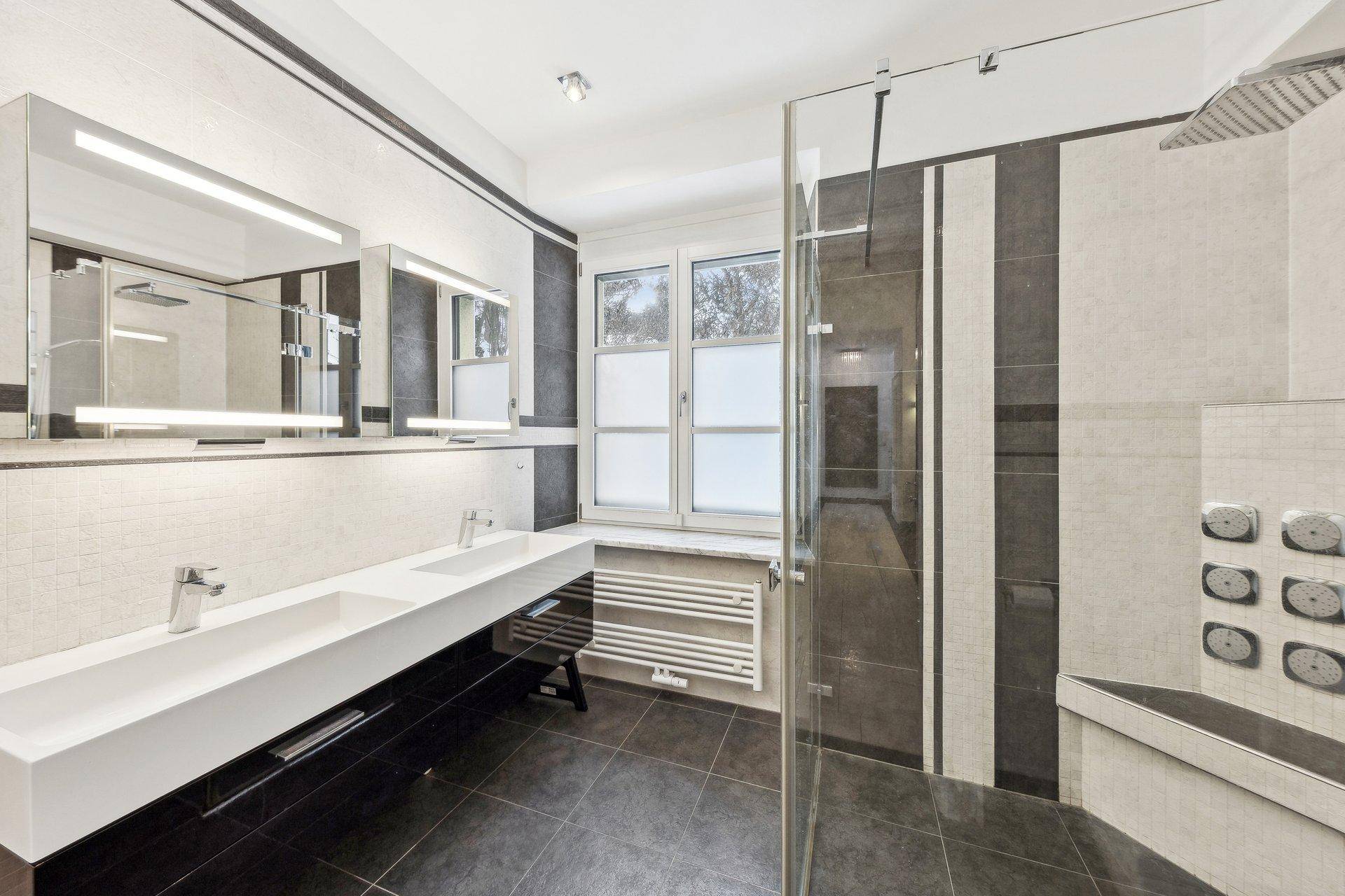 Modernes Badezimmer mit Doppelwaschbecken, großer Dusche und dunklen Fliesen.