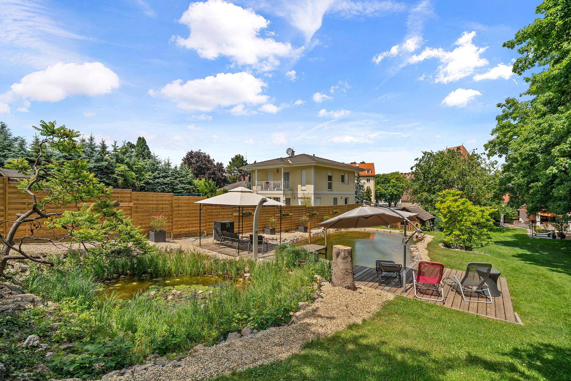Weitläufiger Gartenbereich mit Naturteich, Holzdeck und verschiedenen Sitzgelegenheiten im Freien.