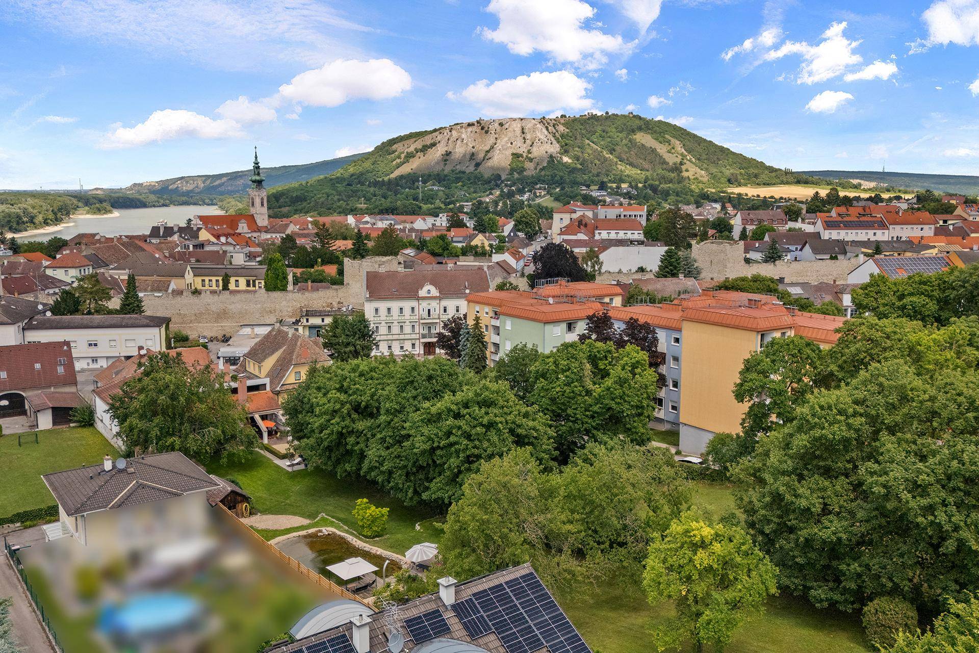 Weitläufige Luftaufnahme einer Stadt am Fluss mit Bergkulisse und grünen Dächern.