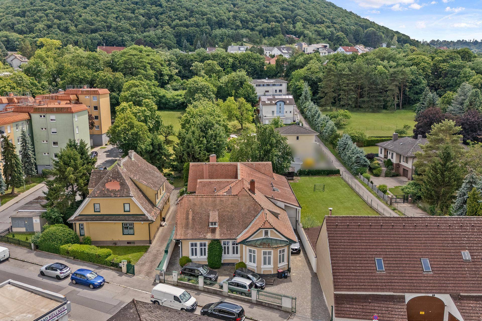 Luftaufnahme einer Wohnsiedlung mit Häusern, Gärten und bewaldeten Hügeln im Hintergrund.