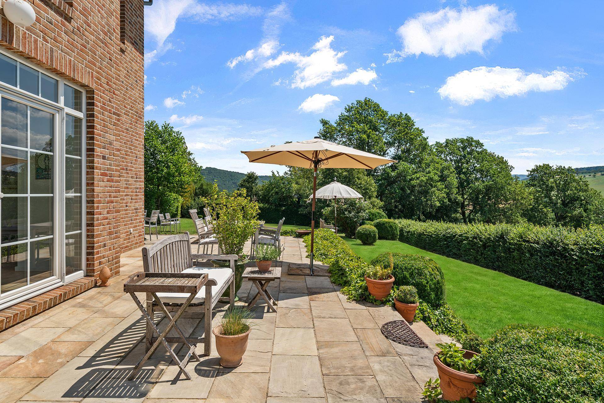 Sonnige Terrasse mit Holzmöbeln und einem Sonnenschirm, umgeben von grüner Hecke und Bäumen.