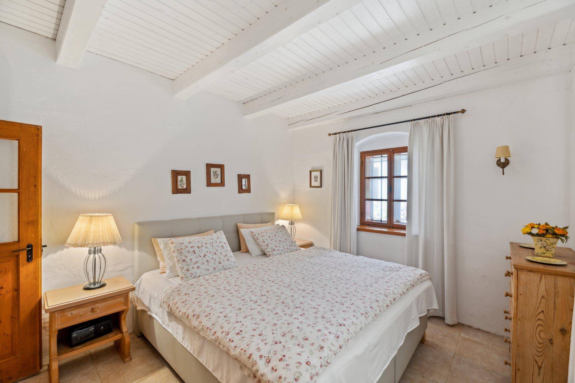 Helles Schlafzimmer mit weiß gestrichenen Holzbalken, Doppelbett und rustikalem Holzboden.