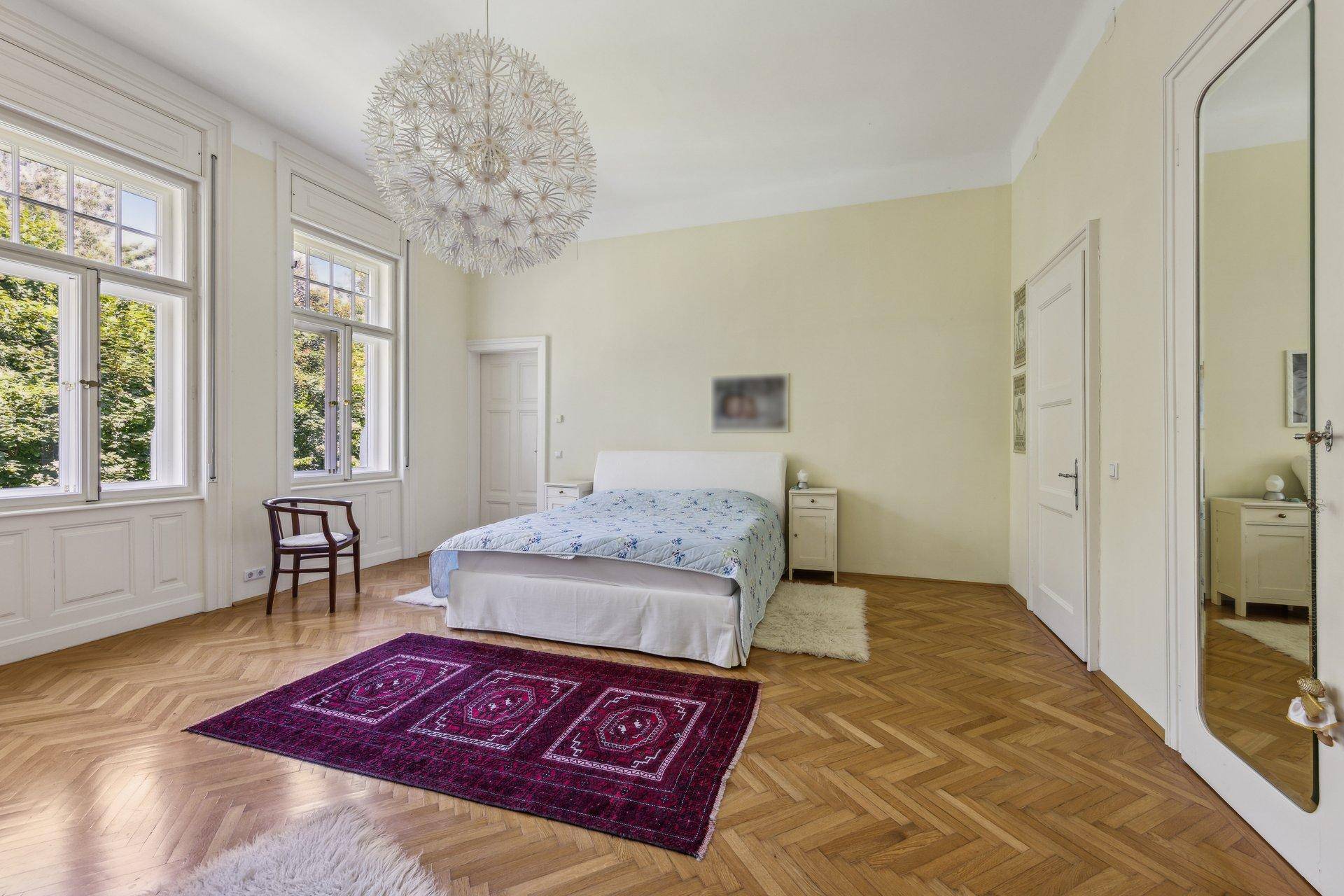 Helles Schlafzimmer mit Parkettboden, hohen Decken und großen Fenstern mit Blick ins Grüne.