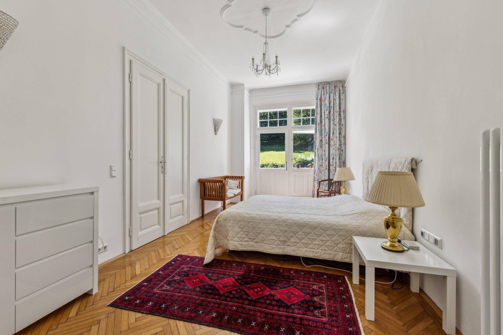 Gemütliches Schlafzimmer mit Parkettboden, hohen Decken und Blick auf den Garten.