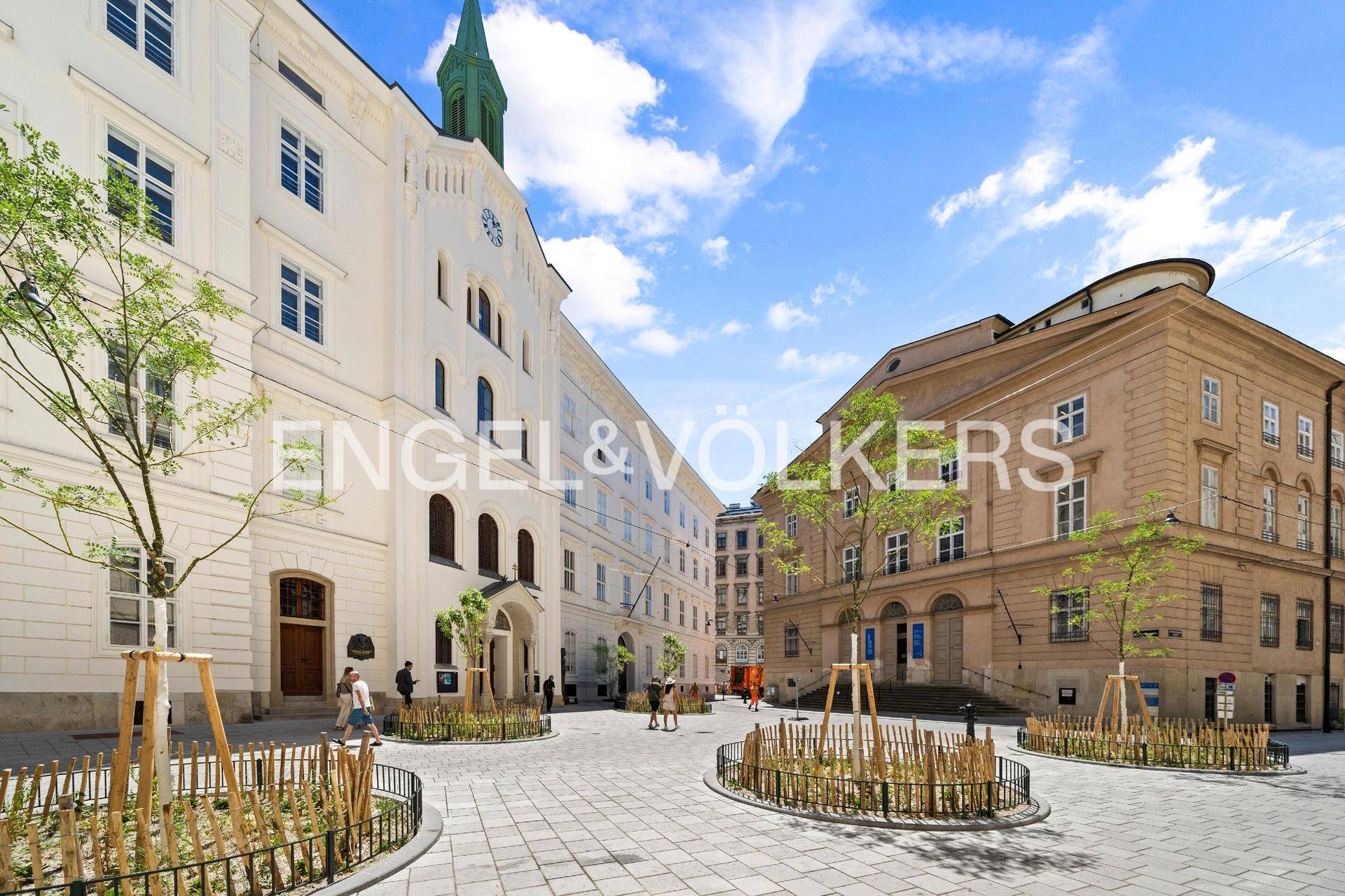 Historische Stadtansicht mit gepflastertem Platz, eleganten Gebäuden und jungen Bäumen unter blauem Himmel.
