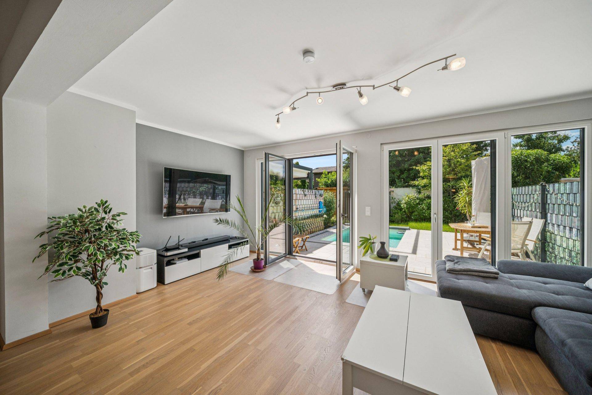 Geräumiges Wohnzimmer mit Wandfernseher, Holzboden und großen Fenstern zum Außenbereich mit Pool.