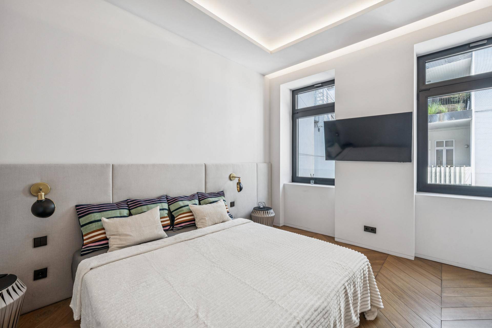 Schlafzimmer mit bequemem Bett, gepolstertem Kopfteil und zwei Fenstern.