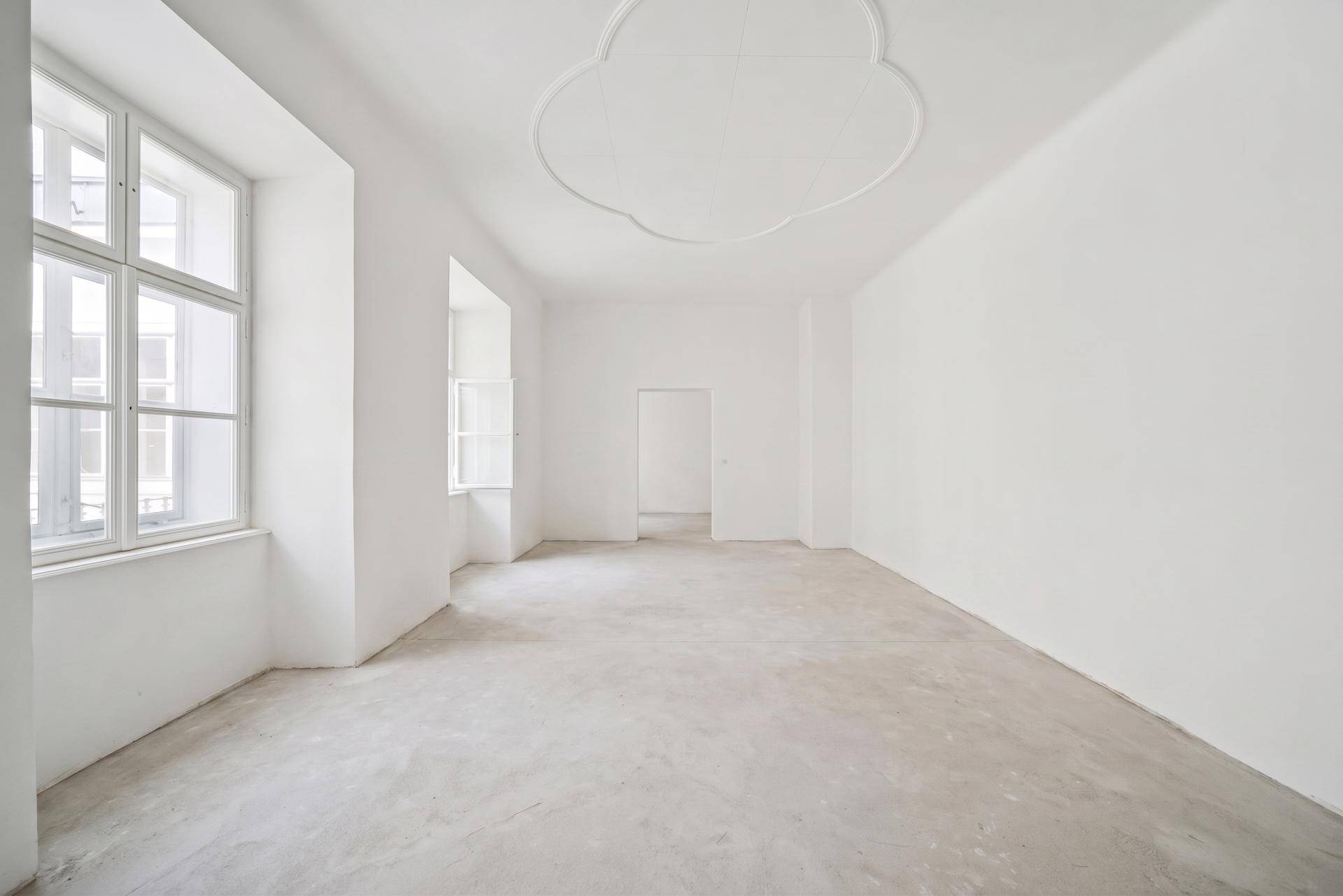 Geräumiger, heller Raum mit Betonboden, hohen Fenstern und dekorativer Stuckdecke.