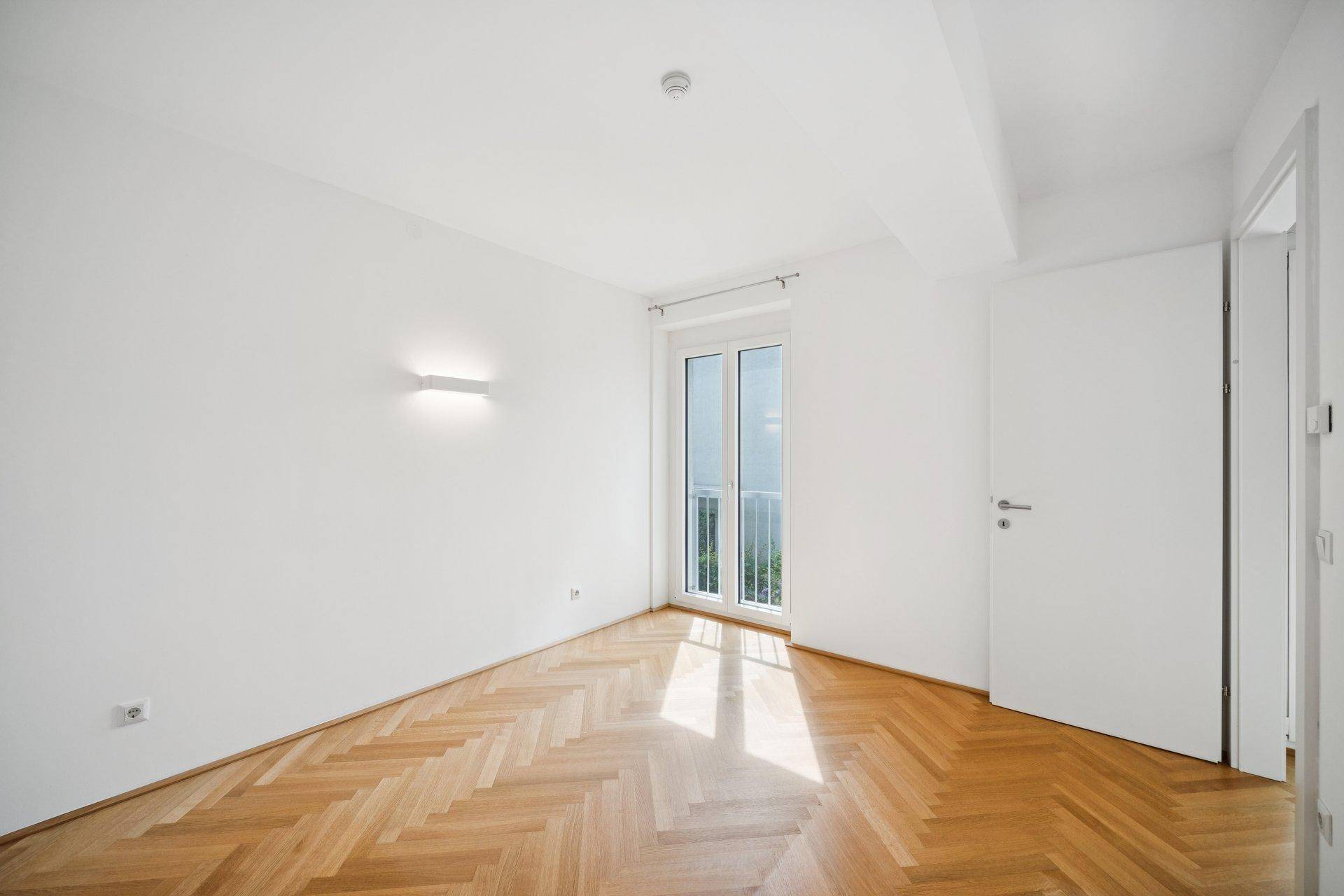 Helles Schlafzimmer mit Fischgrätparkett und Zugang zu einem französischen Balkon.