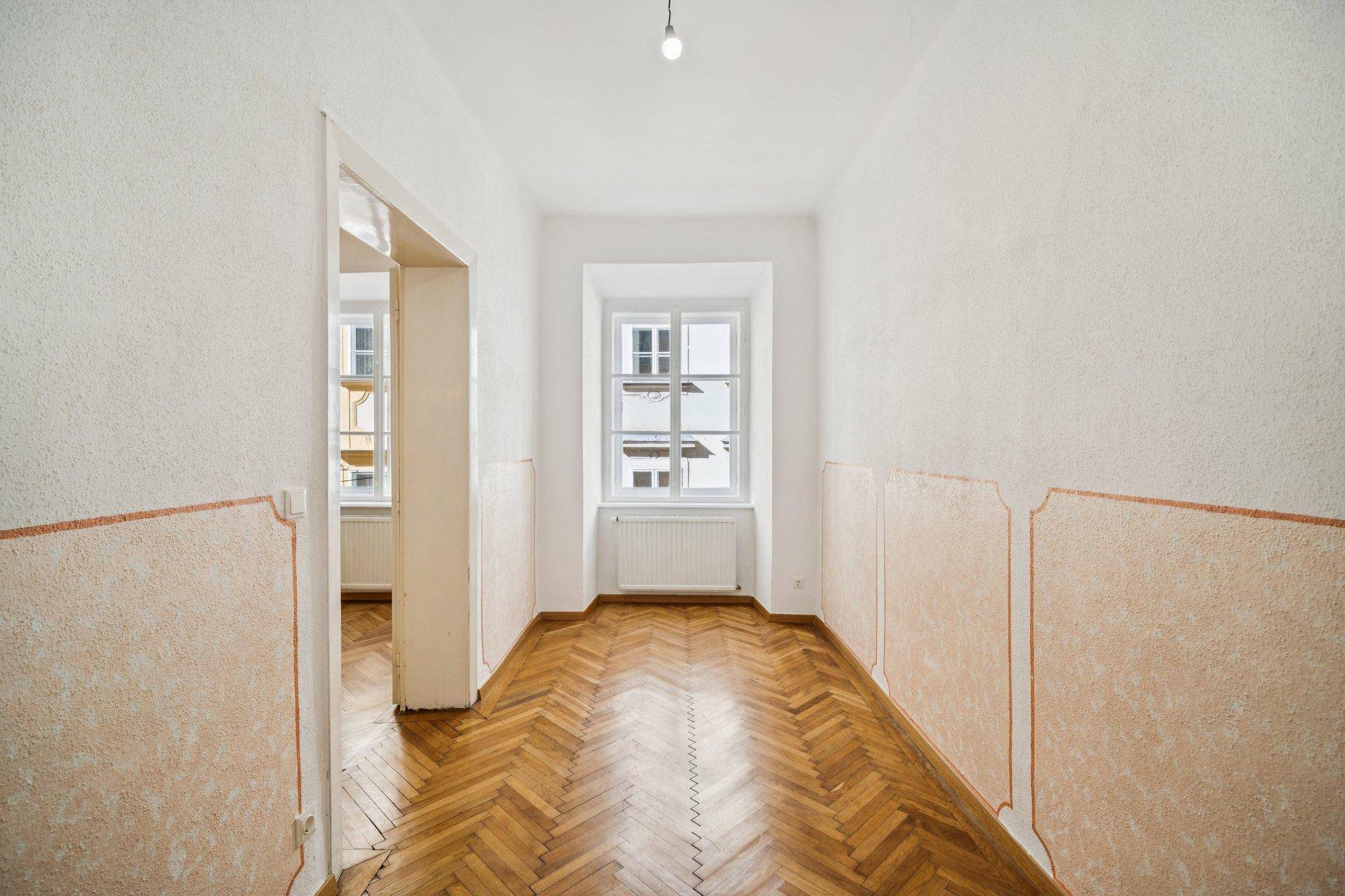 Langer Flur mit Fischgrätparkett, einem Fenster am Ende und Wänden in hellem Pastellton.