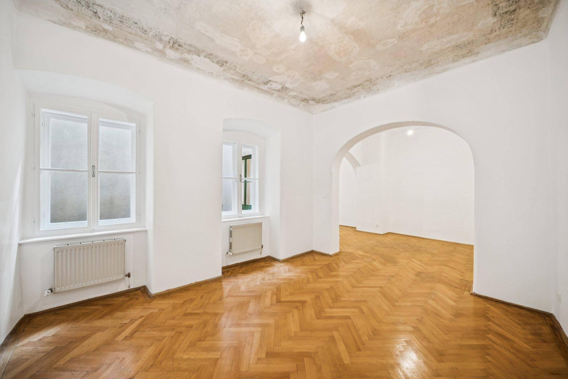 Geräumiges Zimmer mit Fischgrätparkett, zwei Fenstern und einem Bogen zu einem weiteren Bereich.