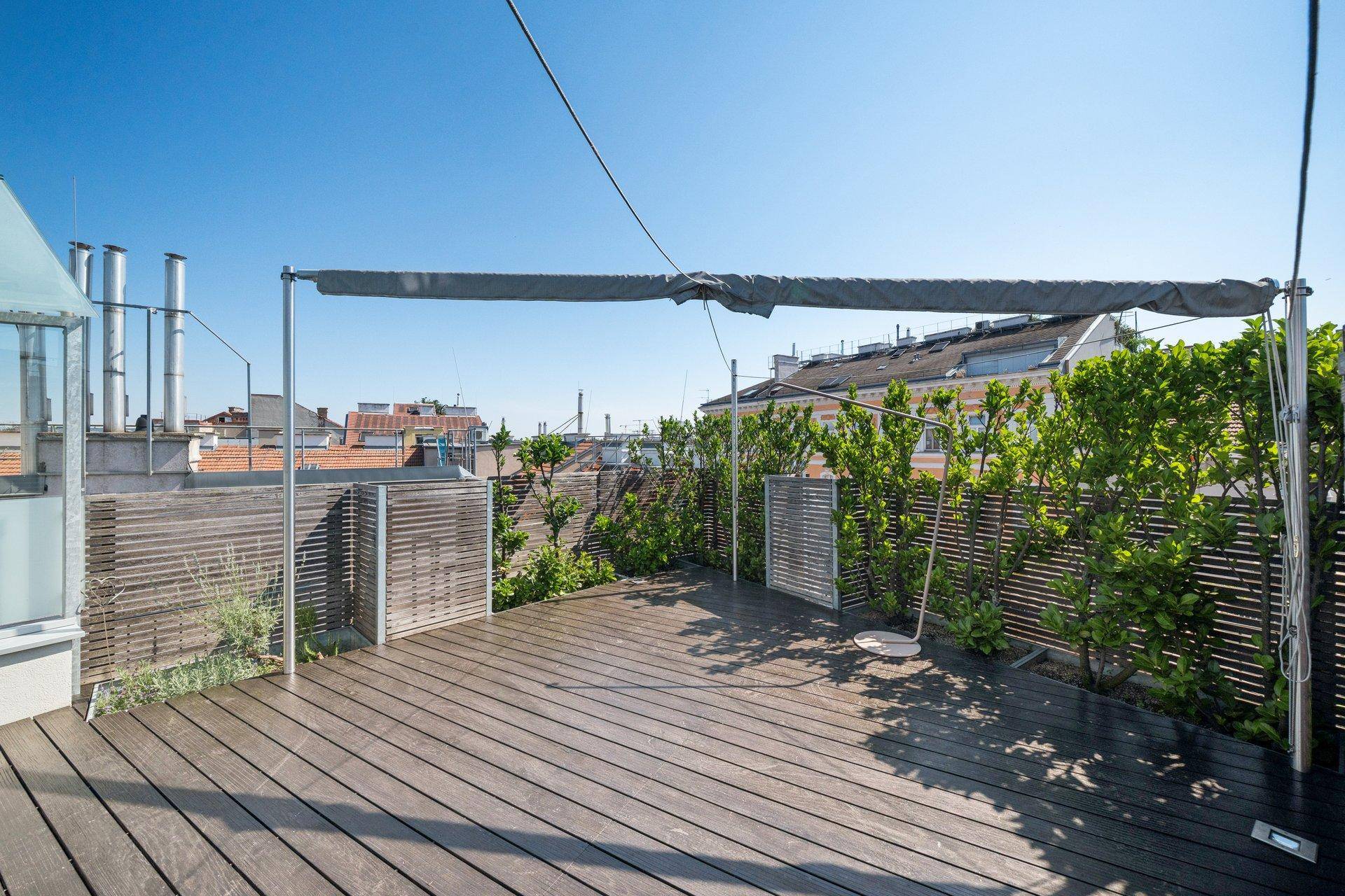 Geräumige Dachterrasse mit Holzdeck, Sichtschutz und viel Grün unter blauem Himmel.