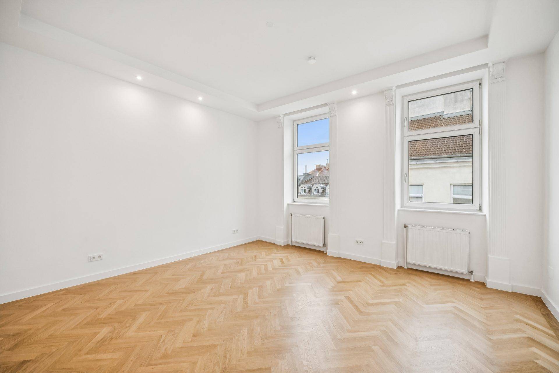 Geräumiges Zimmer mit Fischgrätparkett und zwei Fenstern, das viel Tageslicht hereinlässt.
