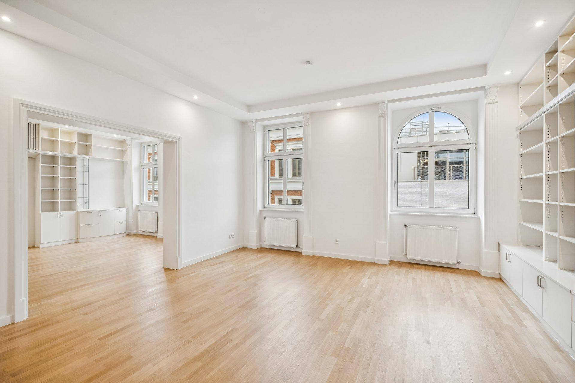 Helles Wohnzimmer mit großen Fenstern, Parkettboden und Zugang zu einem weiteren Raum mit Einbauregalen.