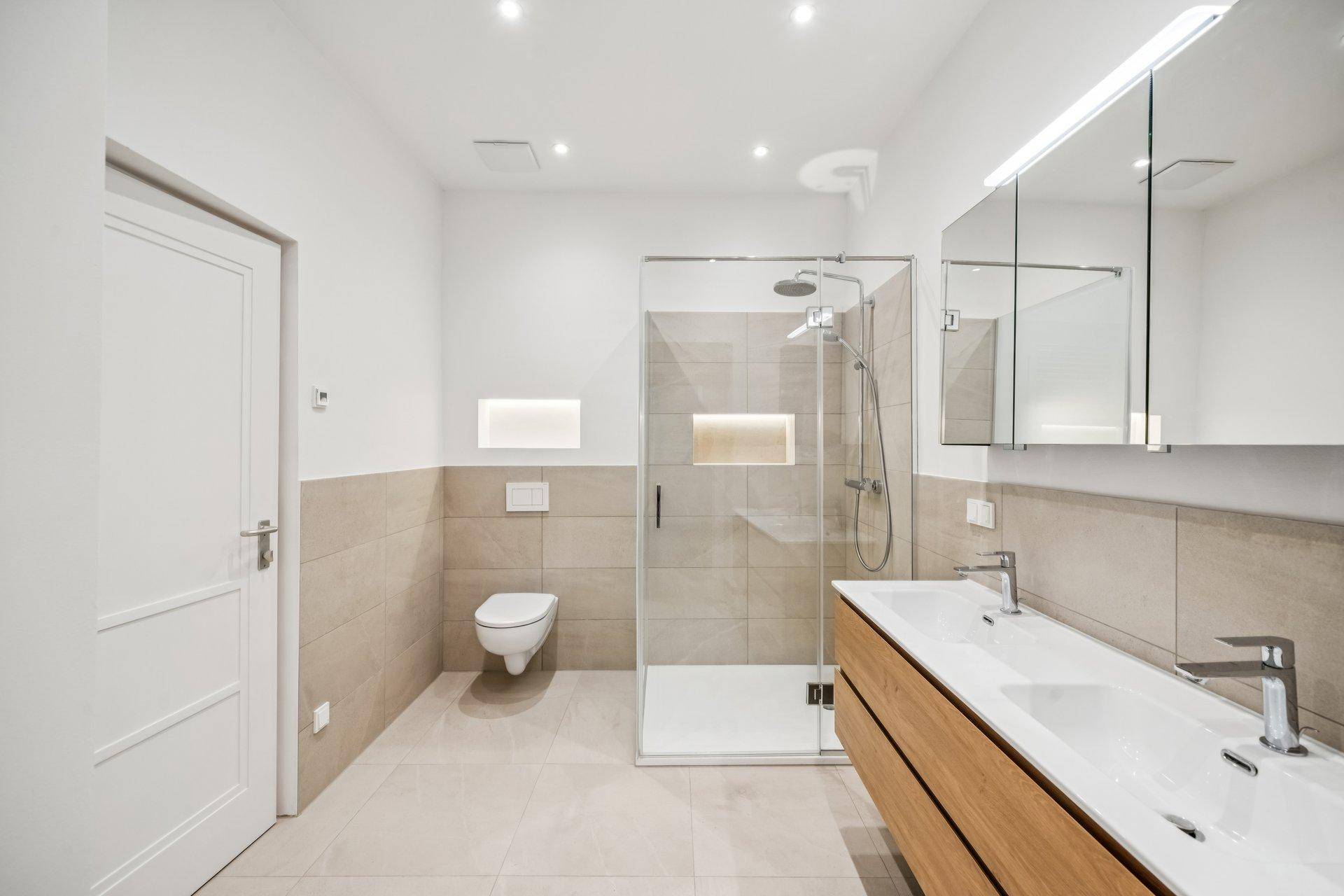 Modernes Badezimmer mit Doppelwaschbecken, begehbarer Dusche und Wand-WC in hellen Farbtönen.