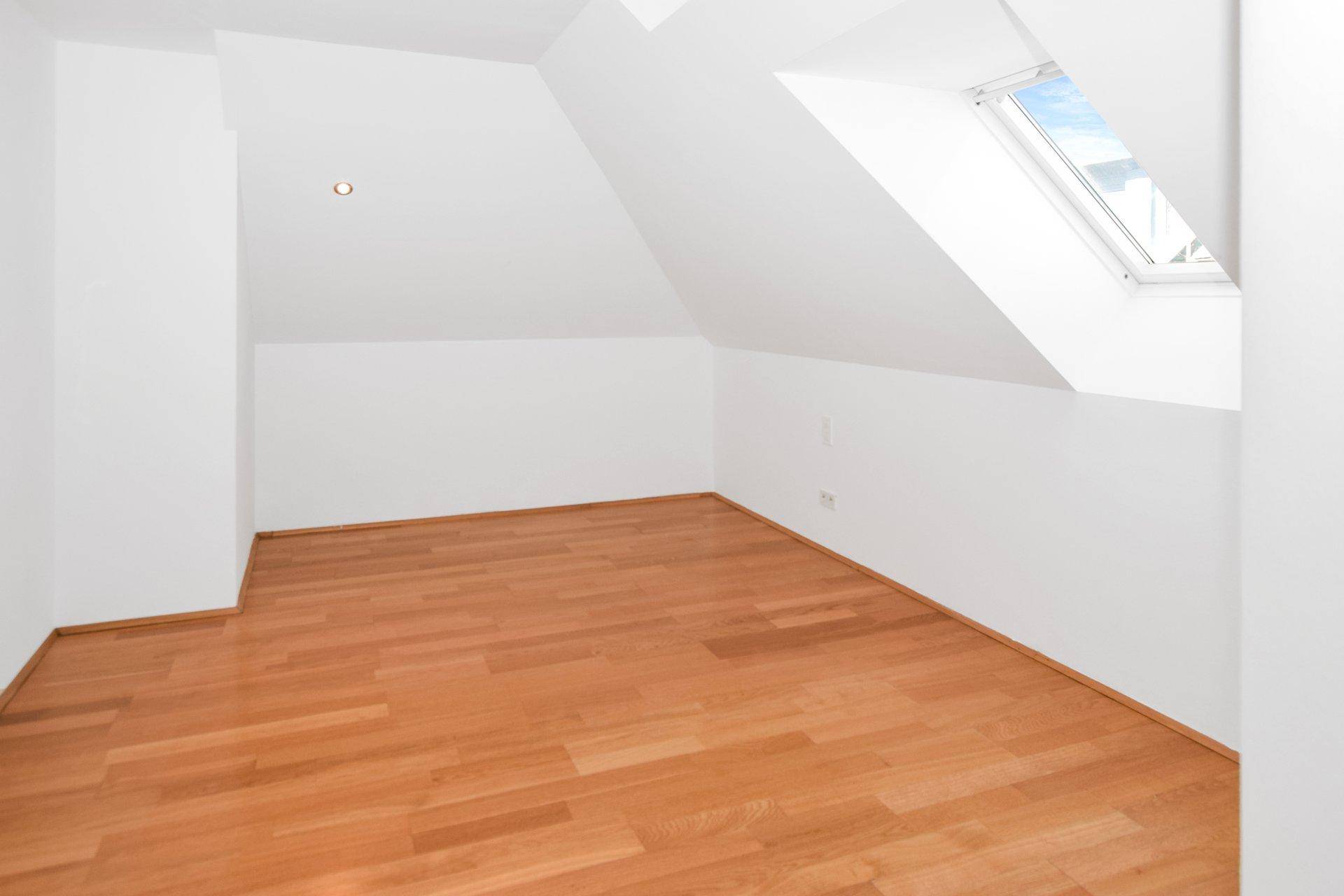 Helles Zimmer im Dachgeschoss mit Holzfußboden und einem Dachfenster, das den Raum mit Licht füllt.