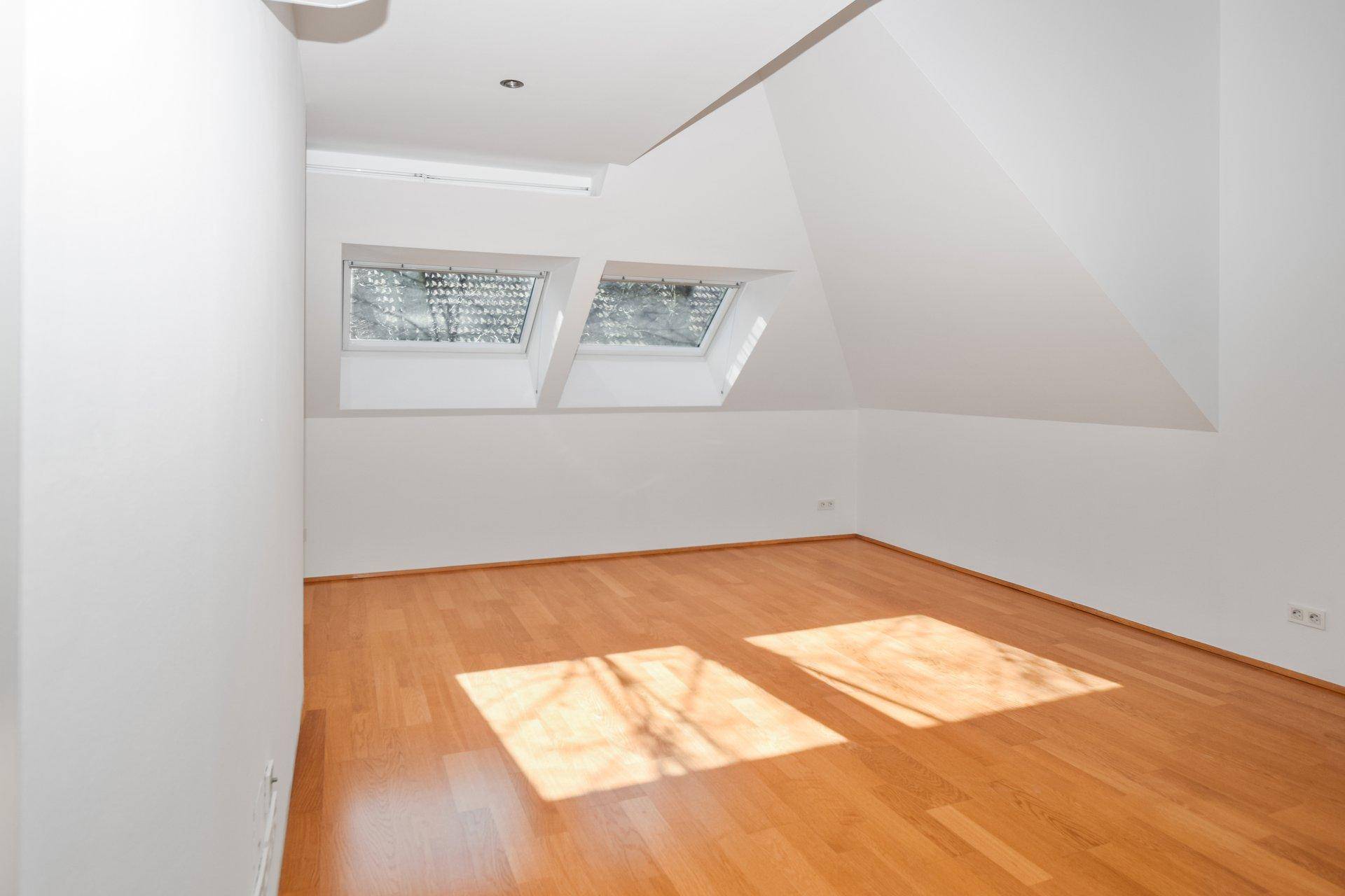 Helles Zimmer im Dachgeschoss mit Holzfußboden und zwei Dachfenstern, die viel Sonnenlicht hereinlassen.